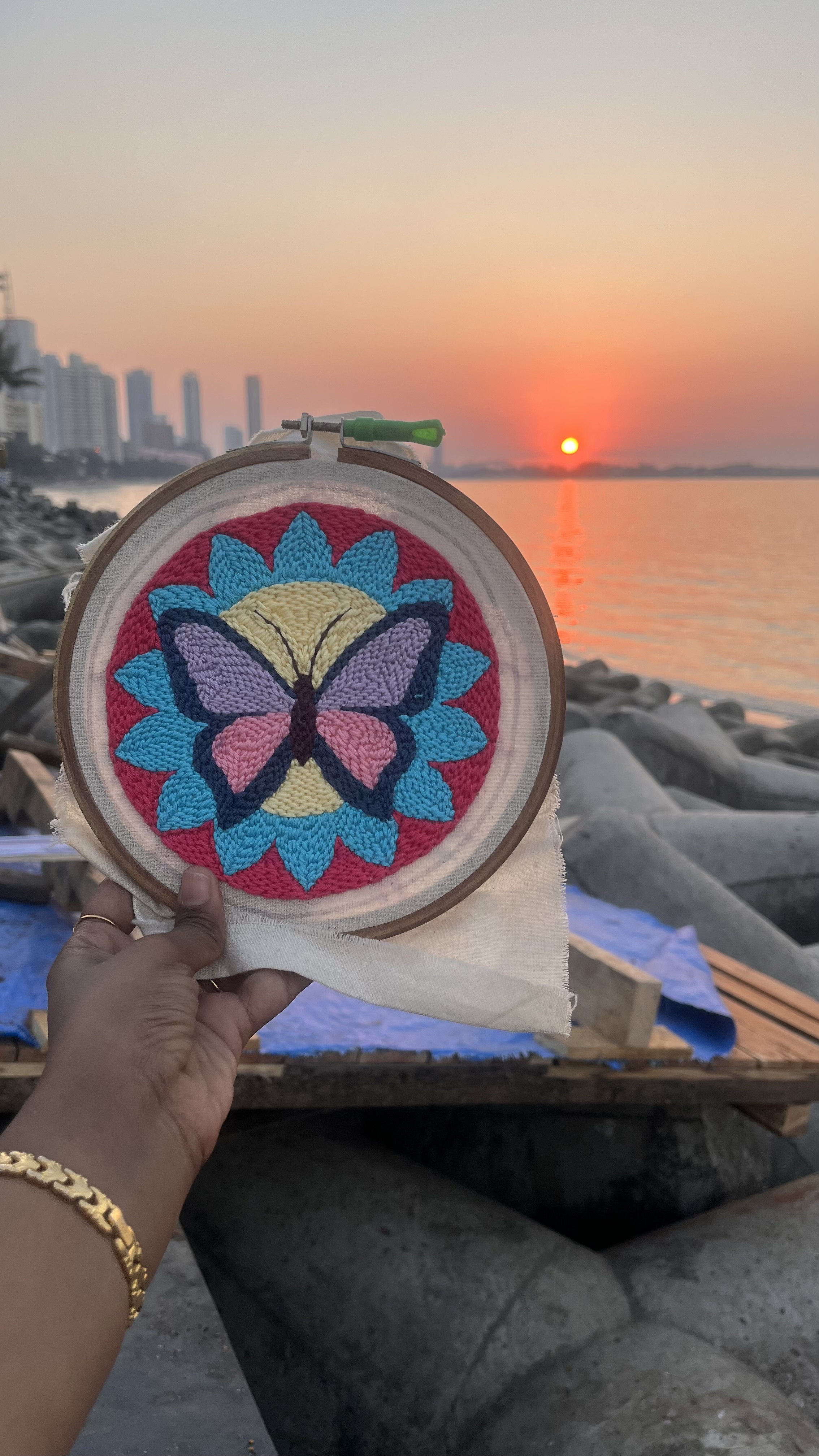 Finished butterfly punch needle hoop held up against a Marine Drive sunset, colourful and warm